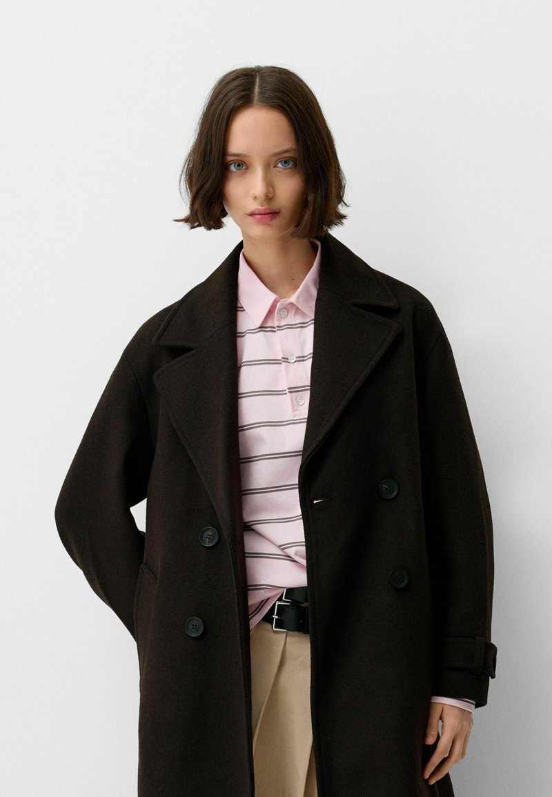 Ciemnobrązowy płaszcz dwurzędowy z dużymi guzikami nałożony na jasnoróżową polówkę w paski. Prosty design z luźnym krojem i klapami.