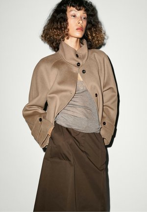 Frau mit lockigem Haar, die eine beige, geknöpfte Jacke, einen grauen Oberteil und einen dunkelbraunen Rock trägt und vor einem einfarbigen hellen Hintergrund steht.
