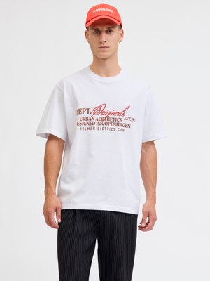 Άνδρας που φοράει λευκό γραφικό T-shirt με κόκκινο κείμενο, μαύρο παντελόνι με ρίγες και κόκκινο καπέλο με την επιγραφή "originals.1990" επί απλής λευκής φόντο.