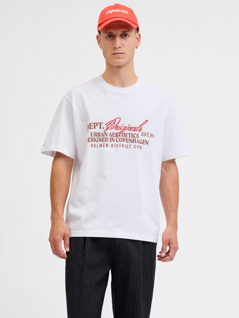 Mann trägt ein weißes T-Shirt mit rotem Schriftzug, schwarze Nadelstreifenhose und eine rote Kappe mit der Aufschrift "originals.1990" vor weißem Hintergrund.