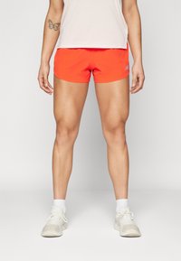 Shorts de sport orange vif avec une texture lisse, dotés d'une taille élastique et d'un détail de logo sur les côtés, associés à des sneakers gris clair.