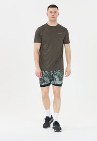 Nuori mies seisoo ja katsoo eteenpäin, pukeutuneena ruskeaan lyhythihaiseen paitaan, vihreisiin kuviollisiin shortsit, valkoisiin sukkiin ja mustiin juoksukenkiin.