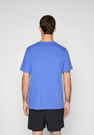 Mężczyzna ubrany w jednokolorowy niebieski T-shirt z krótkim rękawem i czarne szorty, stoi tyłem na jednolitym jasnym tle.