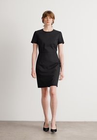 Black fitted dress with short sleeves, round neckline, and subtle texture; paired with black high heels; clean, minimal design.