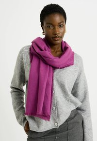 Bufanda púrpura colgada alrededor del cuello, hecha de un tejido suave, con bordes con flecos. Llevada sobre un cárdigan gris con sutiles adornos.
