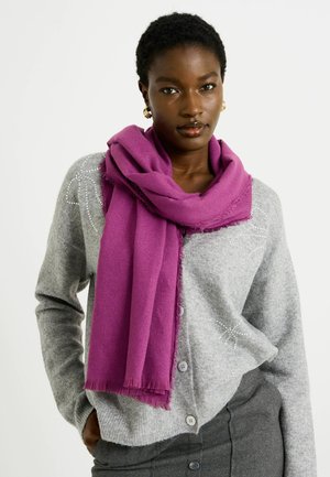 Bufanda púrpura colgada alrededor del cuello, hecha de un tejido suave, con bordes con flecos. Llevada sobre un cárdigan gris con sutiles adornos.