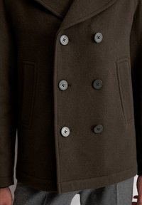 Podwójnie zapinany płaszcz-peacoat w ciemno-brązowej wełnie z kieszenią na piersi, dużymi szarymi guzikami i wyrazistymi klapami, charakteryzujący się gładkim, teksturowanym wykończeniem.