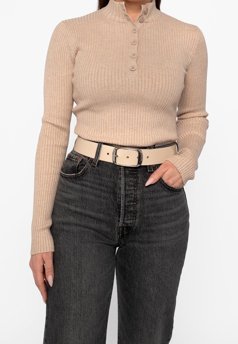 Femme portant un haut beige côtelé à manches longues avec des boutons et un jean noir taille haute avec une ceinture beige.