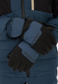 Osoba s modro-černými zateplenými rukavicemi přes modrou bundu s polstrováním, držící si ruce zkřížené na pase.