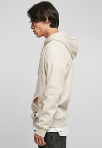 Beige hettegenser laget av myk stoff, med en front kangaroo-lomme, snorhette, og ribbestrikkede mansjetter. Brukt over en hvit skjorte.