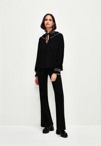 Schwarze Bluse mit Rüschensaum und dekorativem Besatz, kombiniert mit schwarzen, ausgestellten Hosen. Glatter Stoff mit Faltendetails und schwarzen Plateau-Schuhen.
