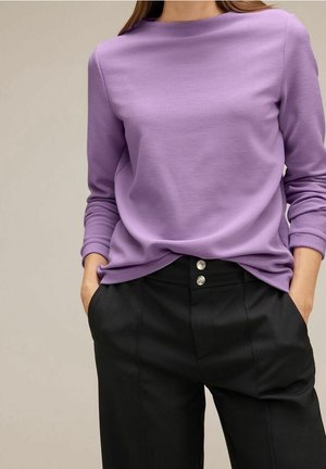 Femme portant un haut côtelé lavande à manches longues, rentré dans un pantalon noir taille haute avec deux boutons argentés, les mains dans les poches.