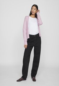 Cardigan rosa chiaro, t-shirt bianca e pantaloni neri morbidi. Scarpe marroni con lacci. Texture morbide, design casual.