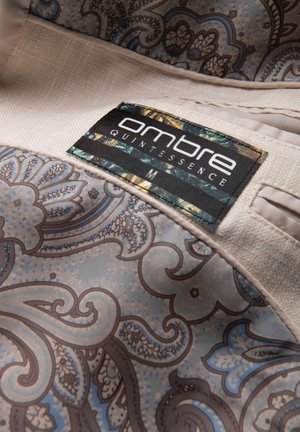 Primer plano de tela beige con estampado paisley azul y marrón y etiqueta cosida que dice "ombre QUINTESSENCE M."