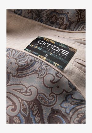 Primer plano de tela beige con estampado paisley azul y marrón y etiqueta cosida que dice "ombre QUINTESSENCE M."
