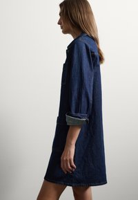 Denim jurk in donkerblauw, met een knopenlijst aan de voorkant, opgerolde mouwen en een borstzak. Glad textuur en een relaxte pasvorm.