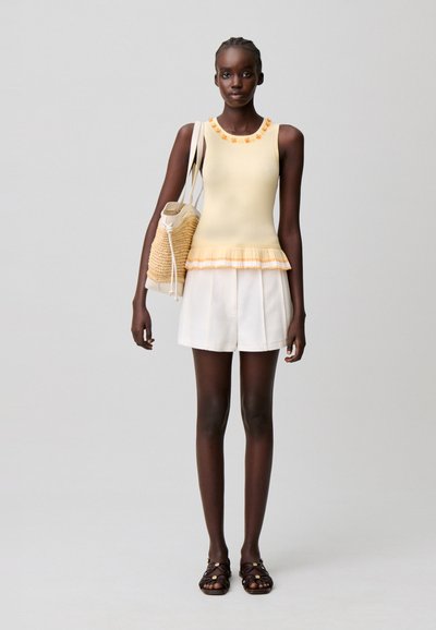 Mujer con una camiseta sin mangas amarilla con ribete floral, pantalones cortos blancos, sandalias oscuras, llevando un bolso tote tejido beige sobre un fondo gris liso.