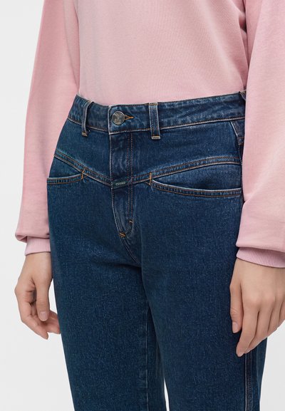Personne portant un jean denim bleu foncé avec des poches avant diagonales uniques et un haut rose à manches longues, mains détendues sur les côtés.