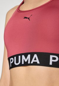 Ružičasti sportski grudnjak s okruglim izrezom, koji sadrži crni Puma logo i široku crnu traku s izrazitim bijelim tekstom "PUMA". Glatka tekstura.