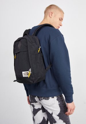 Un homme portant un sweat-shirt bleu marine et un pantalon à motifs noirs et blancs porte un sac à dos noir North Face avec des tirettes de fermeture éclair jaunes.