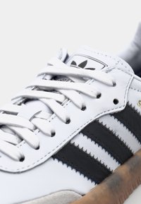 White Adidas sneaker with black stripes, white laces, and gum sole, shown in close-up on a light background.