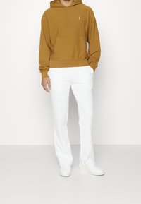 Felpa con cappuccio in pile marrone con tasca frontale e laccio bianco, abbinata a pantaloni bianchi a gamba stretta. Presenta dettaglio logo bianco sui pantaloni.