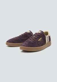 Zapatillas de ante moradas con un diseño de caña baja, que cuentan con franjas crema y detalles dorados, y una suela de goma marrón.