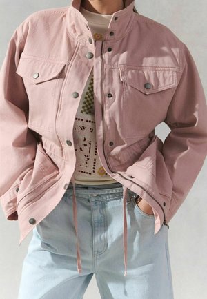 Personne portant une veste rose clair boutonnée par-dessus un pull à motifs et un jean bleu clair, les mains dans les poches.