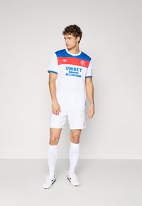 Vīrietis baltā futbola tērpā ar sarkaniem un ziliem akcentiem, stāv un skatās uz sāniem pret fona gaismu.