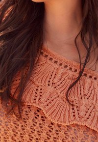 Nærbillede af en person iført en orange strikket sweater med en flæset halsudskæring og langt mørkt hår, der delvist dækker tøjet.