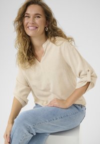 Béžová ľanová blúzka s výstrihom do V a zrolovanými rukávmi, kombinovaná so svetlomodrými džínsami. Hladká textúra a jemný väzobný vzor.