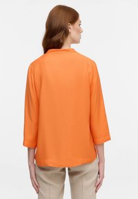 Blusa arancione con maniche a tre quarti, texture liscia e colletto classico, abbinata a pantaloni beige. La vista posteriore mostra i dettagli delle cuciture.