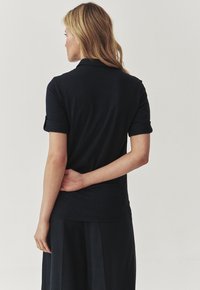 Polo oscuro de color navy con mangas enrolladas, diseño ajustado y textura suave, usado con una falda a juego. La vista trasera muestra un detalle mínimo.