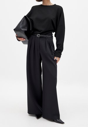 Mujer con pantalones negros de pierna ancha, top negro de manga larga, sosteniendo un gran clutch negro y zapatos negros de punta.