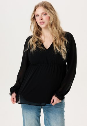 Junge Frau mit langen, welligen blonden Haaren, die eine schwarze Langarmbluse und blaue Jeans trägt, steht vor einem schlichten hellen Hintergrund.
