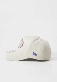 Beige baseball cap med en buet skygge. Har broderet logo i lilla og beige. Indeholder et lille logo akcent på siden.