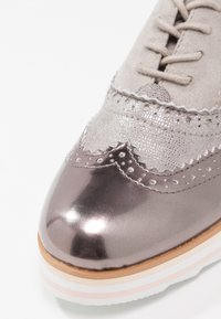Chaussure à lacets en daim gris et métallique avec un design festonné, un bout poli et une semelle à plusieurs couches rayées en blanc, rose et beige.