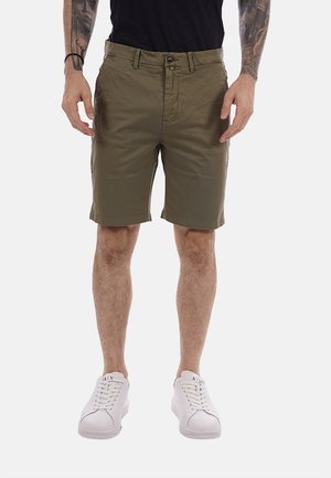 Uomo con pantaloncini verde oliva fino al ginocchio, camicia nera, scarpe da ginnastica bianche, e braccia tatuate con mani rilassate lungo i fianchi.