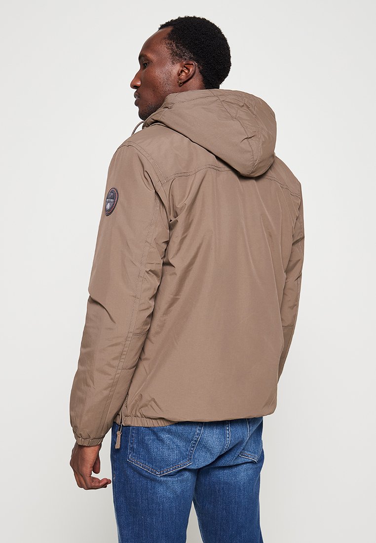 Chaqueta aislante marrón con capucha, que presenta un parche de logo redondo en la manga y puños elásticos, combinada con jeans de mezclilla azul.