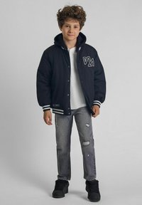 Veste varsity noire avec des rayures blanches, boutons à pression sur le devant et logo ; associée à un jean gris clair déchiré et des baskets noires.