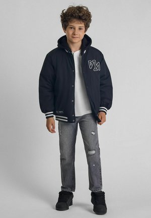 Veste varsity noire avec des rayures blanches, boutons à pression sur le devant et logo ; associée à un jean gris clair déchiré et des baskets noires.
