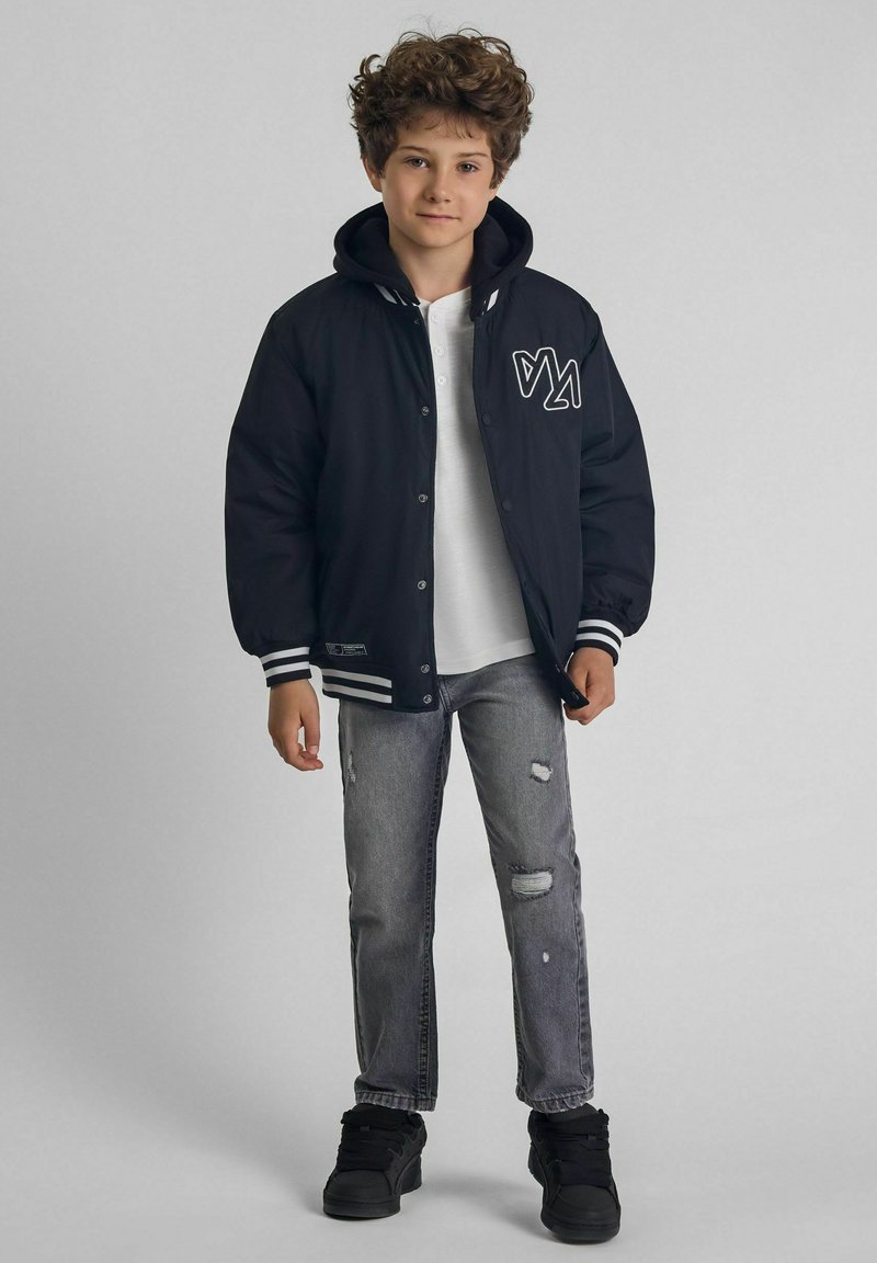 Veste varsity noire avec des rayures blanches, boutons à pression sur le devant et logo ; associée à un jean gris clair déchiré et des baskets noires.