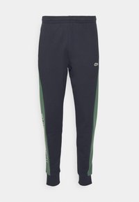 Marina joggers med gröna sidopaneler, elastisk midja och mudd nederkant. Har en logo nära midjan och tryckt text längs sidan.