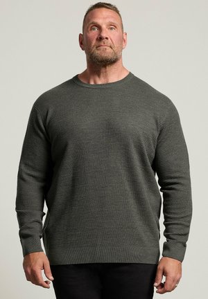 Homme aux cheveux courts et barbe portant un pull texturé gris foncé à manches longues et un pantalon noir, debout devant un fond uni.