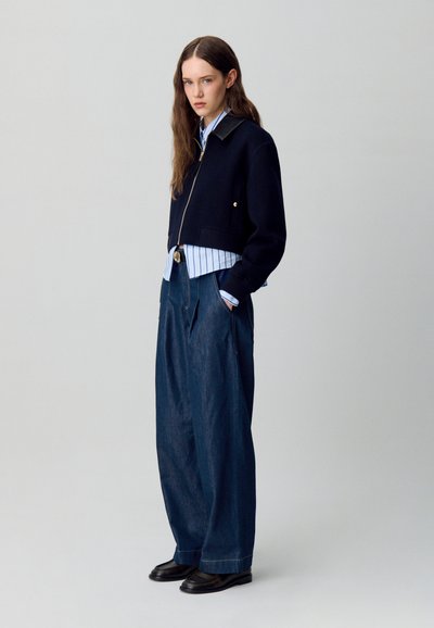 Chaqueta navy corta con cuello de cuero, llevada sobre una camisa de rayas azul clara. Pantalones de mezclilla oscuros y holgados y zapatos negros completan el atuendo.