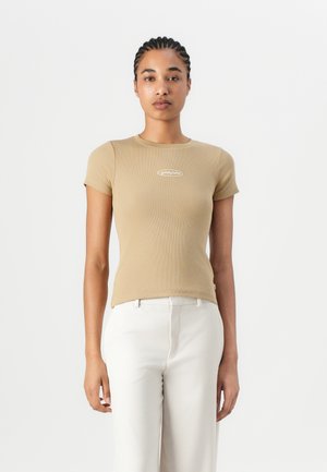 Beige ribbel-T-Shirt mit kurzen Ärmeln und Rundhalsausschnitt, mit einem kleinen Logo in der Mitte. Kombiniert mit weißen, hochtaillierten Hosen.