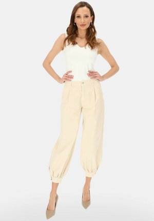 Kobieta stojąca z rękami na biodrach, ubrana w biały bezrękawnik, beżowe szerokie spodnie z wysokim stanem i beżowe szpiczaste szpilki.