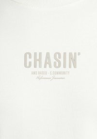 Baltas džemperis su tekstūruotu light grey logotipu, kuriame užrašyta "CHASIN' AMS BASED - C.COMMUNITY" ir parašas apačioje.