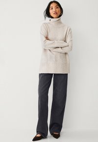 Una donna che indossa un maglione beige a collo alto, jeans larghi scuri, scarpe nere a punta e orecchini a cerchio dorati, in piedi con le braccia incrociate.