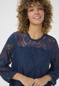 Tamnoplavi top s složenim čipkastim detaljima, s valovitim izrezom i rukavima do tri četvrtine. Izrađen od lagane tkanine.